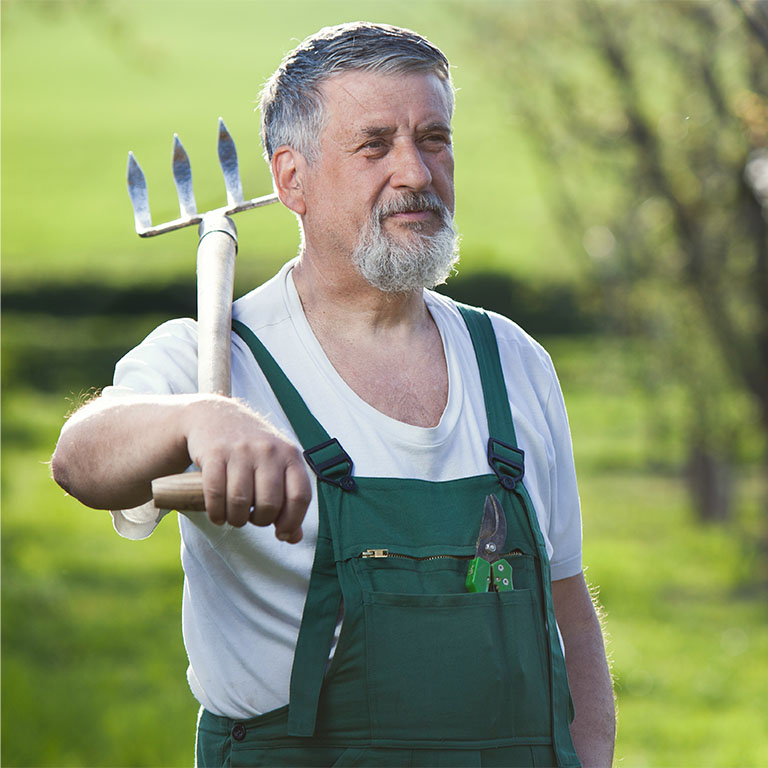 Ein älterer Mann in grüner Arbeitskleidung hält eine dreizinkige Hacke in der Hand.